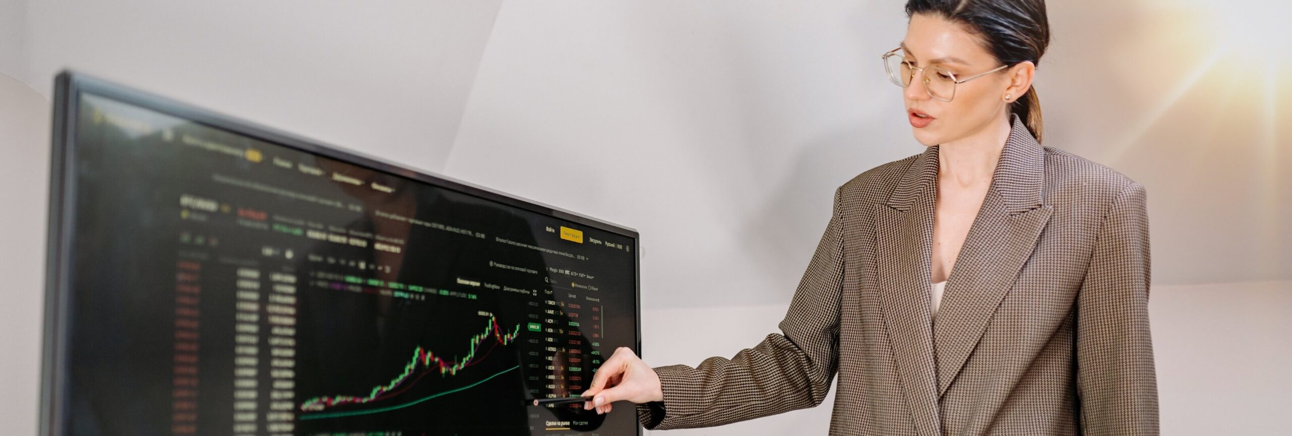 Woman in glasses and a checked blazer pointing at a large screen displaying a candlestick chart and market data tables, representing labor market data for capital markets.